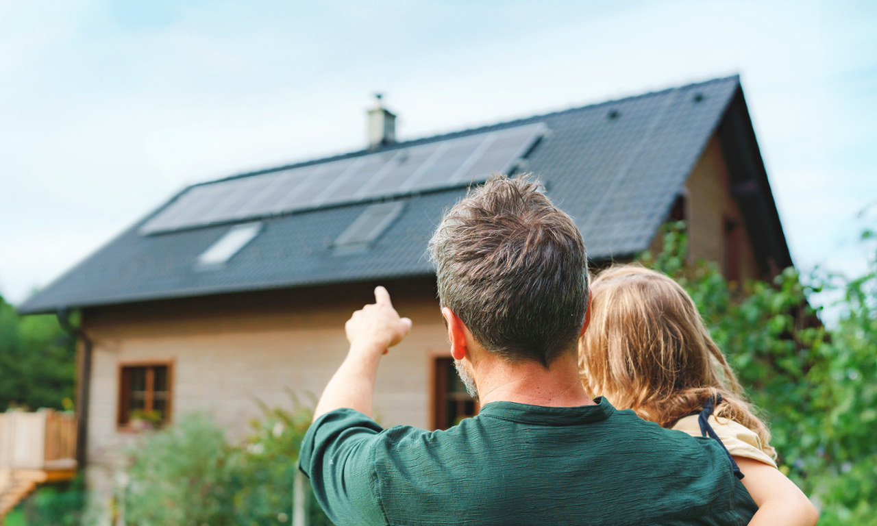 Vater steht mit seiner Tochter auf dem Arm mit dem Rücken zur Kamera und zeigt auf ein Haus mit PV-Modulen auf dem Dach.